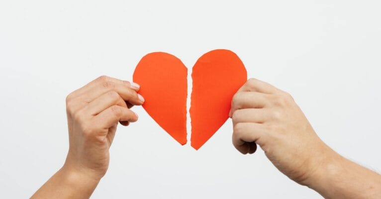 A close-up view of hands holding a torn red paper heart, symbolizing heartbreak or relationship issues.